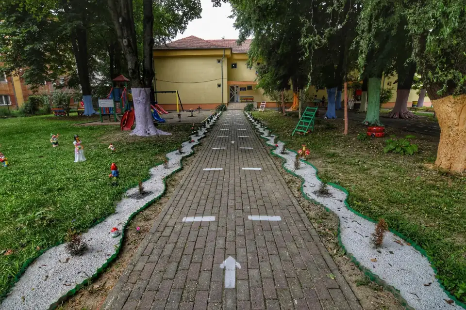 Aleea din curtea grădiniței, mărginită de copaci cu trunchiuri pictate, care duce spre clădirea galbenă