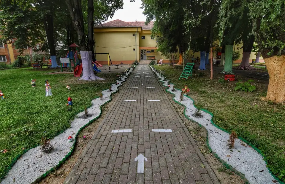Alee pavată spre intrarea grădiniței, mărginită de spiriduși și ciuperci pictate, cu locul de joacă la umbra copacilor